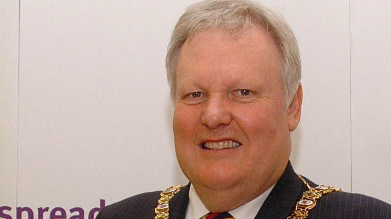 Lord Browne standing smiling at the camera. He is wearing a mayoral chain, a black suit and a tie with red, blue and yellow stripes. 