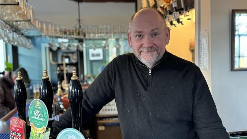 A bald, middle-aged man wearing a black jumper while smiling at the camera. He is stood behind beer pumps behind a bar in a pub