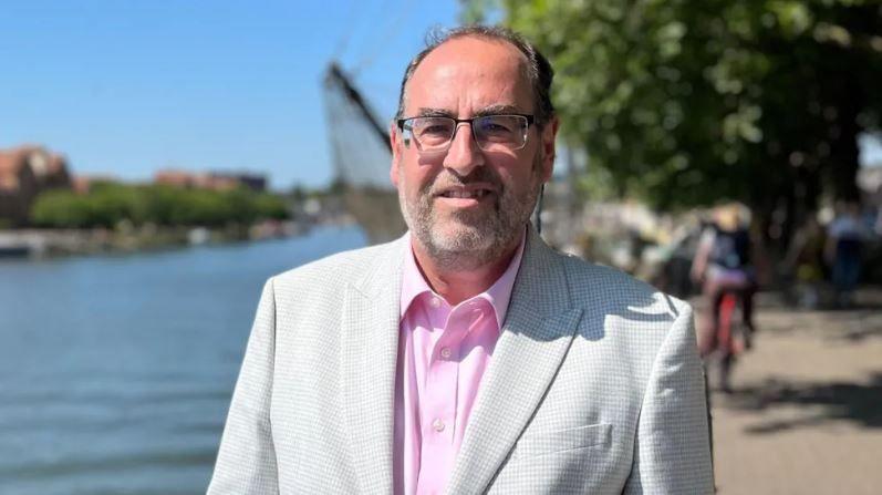 Bristol City Council leader Tony Dyer, wearing glasses and a grey suit with a pink shirt. He is stood in front of a river and a blurred cyclist can be seen behind him.