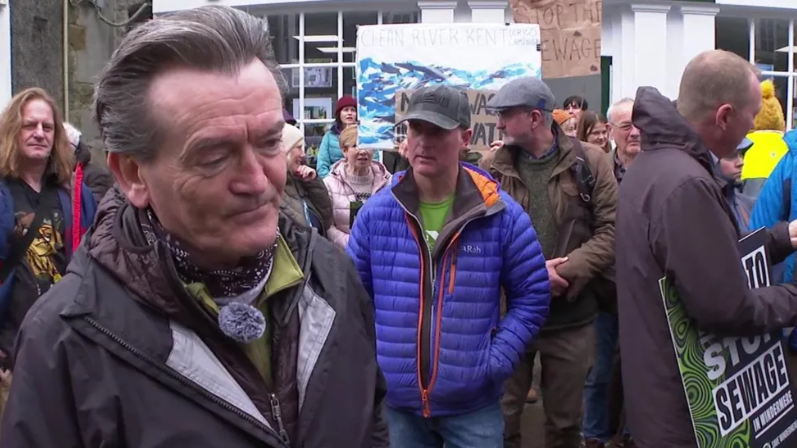 A man with grey hair standing with protestors. Signs read 'stop the sewage'.