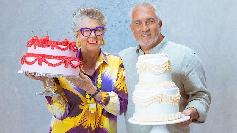 Prue in a yellow and purple shirt with a yellow necklace and purple glasses and Paul in a pale blue shirt both are holding tiered cakes.