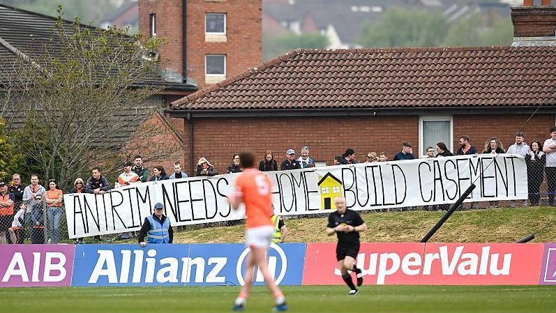 People hold up a sign saying Antrim Needs A Home Build Casement on the side of a pitch. A Gaelic player in orange is seen on the pitch with the referee nearby. There are houses behind the people holding the sign.