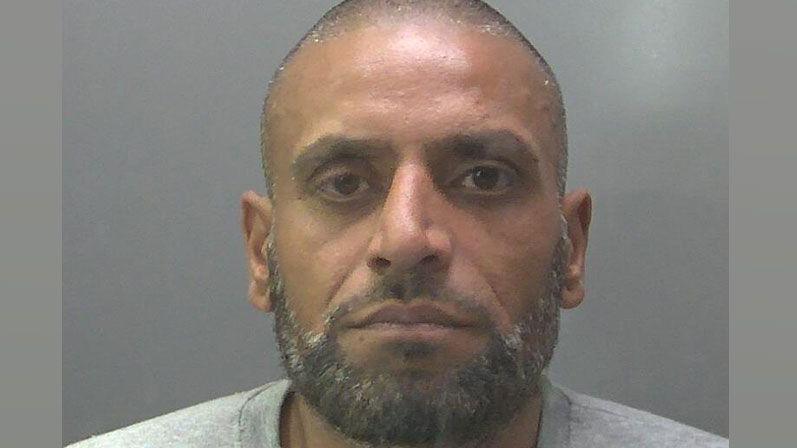 Inam Qureshi in a police mugshot. He has a shaved head showing black and silver stubble and a close cropped black and silver beard. He is wearing a grey sweatshirt.