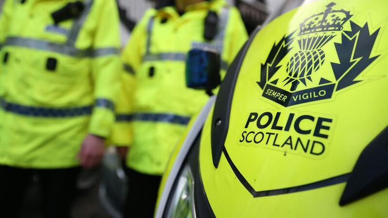 A blurred picture of two Police Scotland officers in fluorescent yellow reflective jackets from the neck down. The front of a motorbike is in the foreground. It bares the crest of Police Scotland which reads Semper Vigilo.