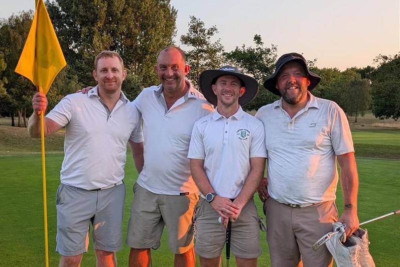 Jonathon Wallace, Mike Ashwin, Ben Read, and Ben Smith pictured at Chipping Sodbury Golf Club, wearing white polo shirts and khaki shorts, standing on the green after completing their16-hour golf challenge with rolling fairways and trees behind them.