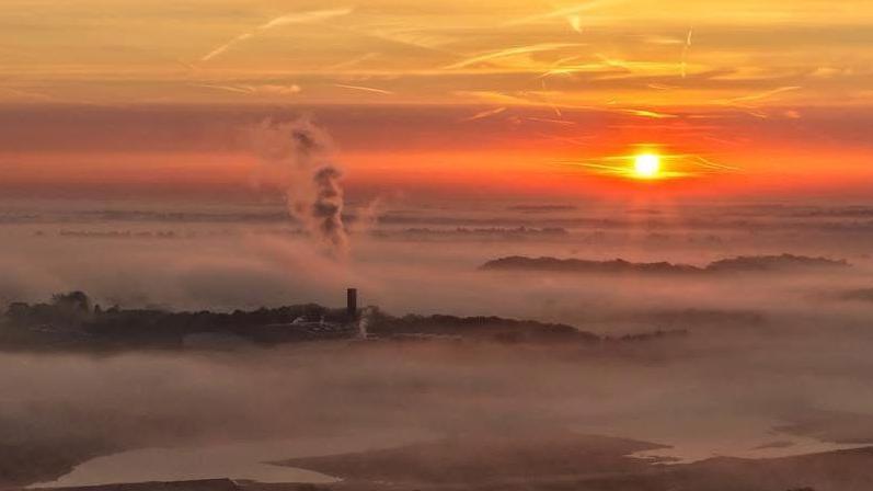 Early morning skyscape with the sun rising above a misty rurual landscape. A chimney stack can be seen with a cloud of steam rising high in the sky from it.