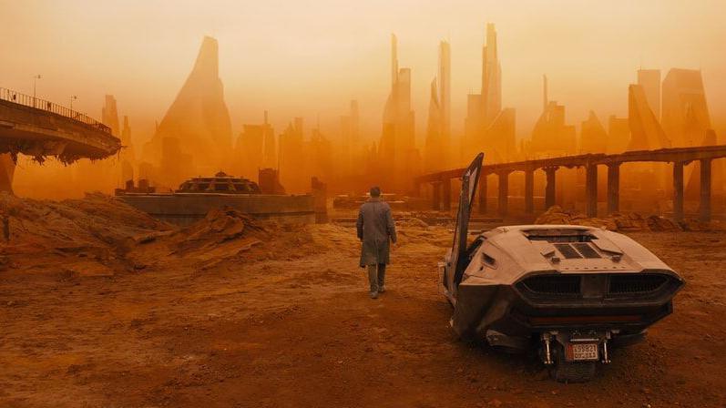 A still image from the film Blade Runner 2049, which shows a vehicle on the right of the image and a person walking towards an orange-hued dystopian-looking city.