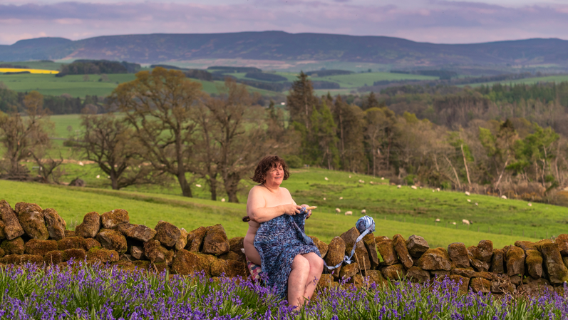 The image shows a woman sitting on a stone wall, with fields and hills behind her. She is naked apart from some strategically-placed knitting covering part of her body 