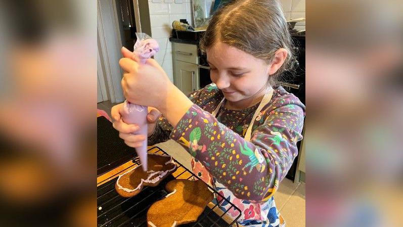 Ivy wearing a colourful top which is brown and green. She is wearing an apron with flowers on it. She is smiling in a kitchen and squeezing an icing bag over two gingerbread cookies on an oven rack. 