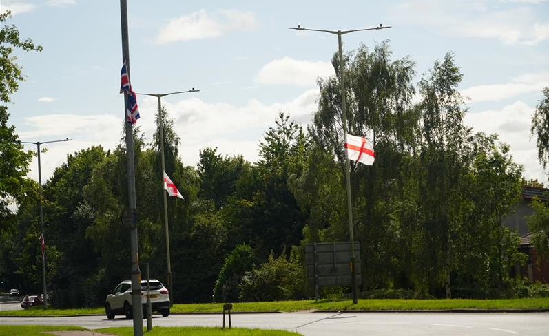St George's Cross flags and a Union flag on lampposts near a roundabout. There is one white car on the road.