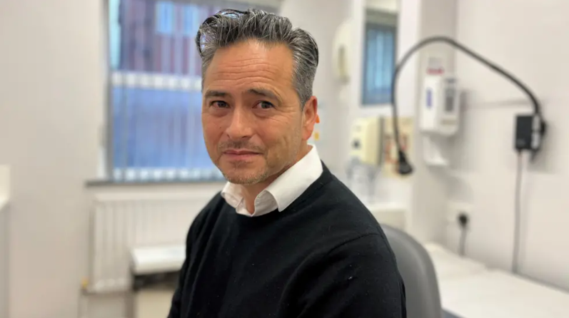 A man with swept-back dark hair and stubble, wearing a white collared shirt and black jumper. He is sitting in a medical consulting room with an examination bed behind him and dispensers on the walls.