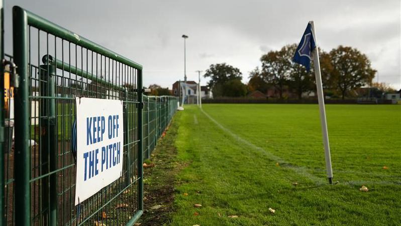 A gate which says: keep off the pitch". A corner flag is in the foreground and the rest of the pitch is in the background