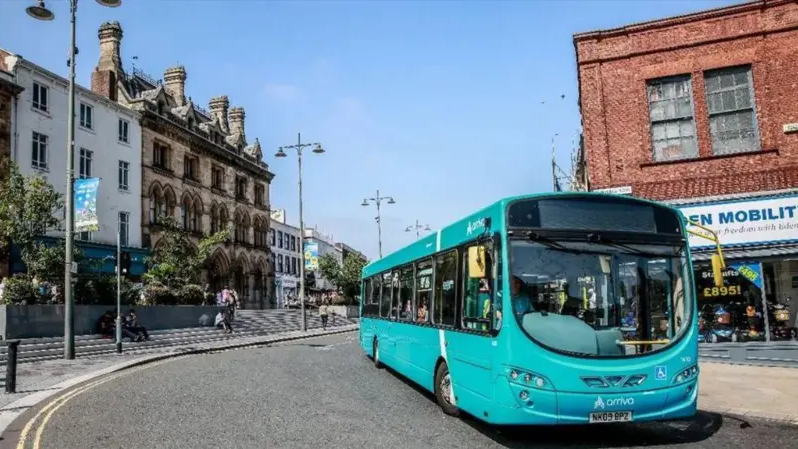 Blue bus on a curved road with buildings on the sides. It is a sunny day. 