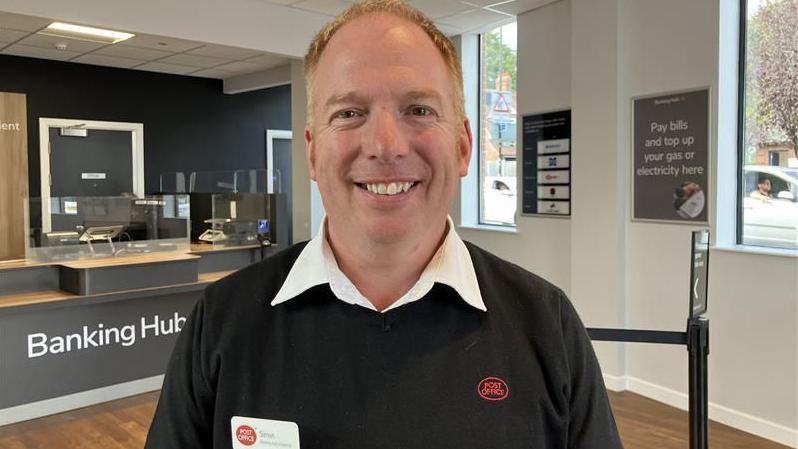 A man with light hair smiling and wearing a white shirt and black jumper with the Post Office logo on the front. In the background is the interior of a bank with a counter and information signs.  