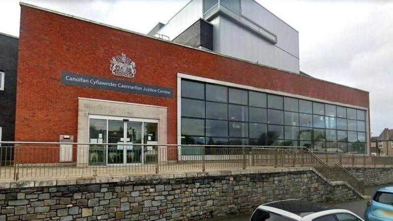 Caernarfon Crown Court has red brick and the court symbol above a four glass door entrance. There is also a large bank of window four panes high and 13 wide covering most of the front of the building. 