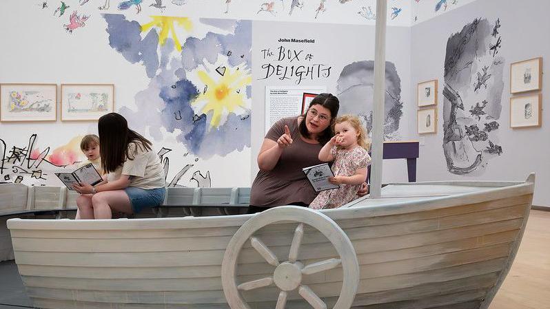 Two women sit in a model boat, each with a young child. They are both reading books to the children. In the background are illustrations on the walls and in frames. 
