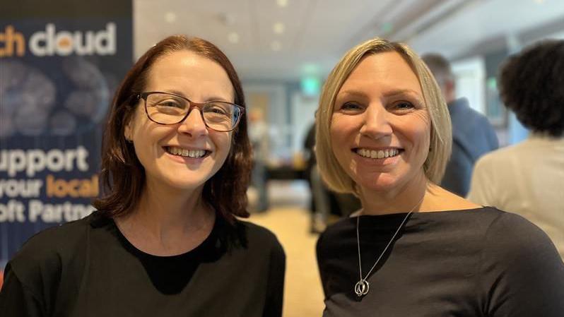 A volunteer and the charity's ceo standing in one of the hotel meeting rooms with a line of volunteers behind them packing boxes. they're both smiling at the camera