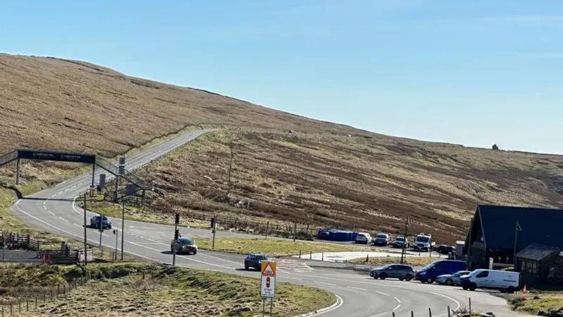 The Mountain Road stretching from the Bungalow through to Brandywell. It is on a sloping mountainside covered in heathland. There is a building at the Bungalow on the right with cars parked in front of it and there are three cars travelling on the road. There is a metal footbridge over the road on the left.