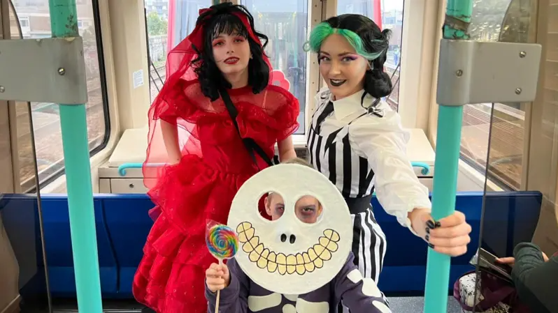 Three young people in detailed costumes - one in pinstriped overalls, another with a circle skeleton mask and another in a ruffled red dress.  