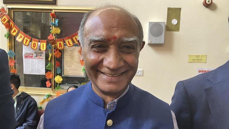 An older man stands inside wearing a dark blue button-up vest with a long-sleeved shirt underneath. He has a red mark painted on his forehead.