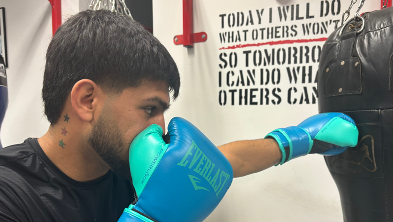 Shows a profile view of Mujeeb jabbing a punch bag with his left hand with a tattoo of three stars - in blue, red and green - on his neck