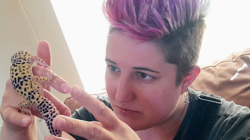 Lianne Abel holding a gecko. Ms Abel has short hair which is long and pink at the top. She has dark eyes and a lip piercing. She is holding a gecko on her right hand. The gecko is beige with black spots. 