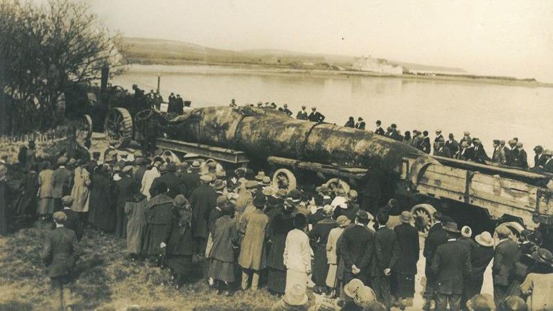 Sepia photograph of crowds of people look on as the dead whale is transported. Derbyhaven can be seen in the background.