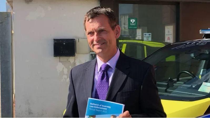 Ruari Hardy stood in front of police vehicles. He is holding a Bailiwick of Guernsey Law Enforcement Annual Report booklet, which is blue. He is wearing a purple shirt, purple spotted tie and black blazer. 