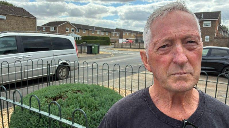 A man is looking directly at the camera. He is outside standing in a residential area. Behind him is a row of houses. 