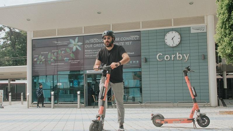 A man with a dark beard wearing a black T-shirt and grey tracksuit trousers rides an orange e-scooter outside Corby railway station. Another orange e-scooter is standing to his left.