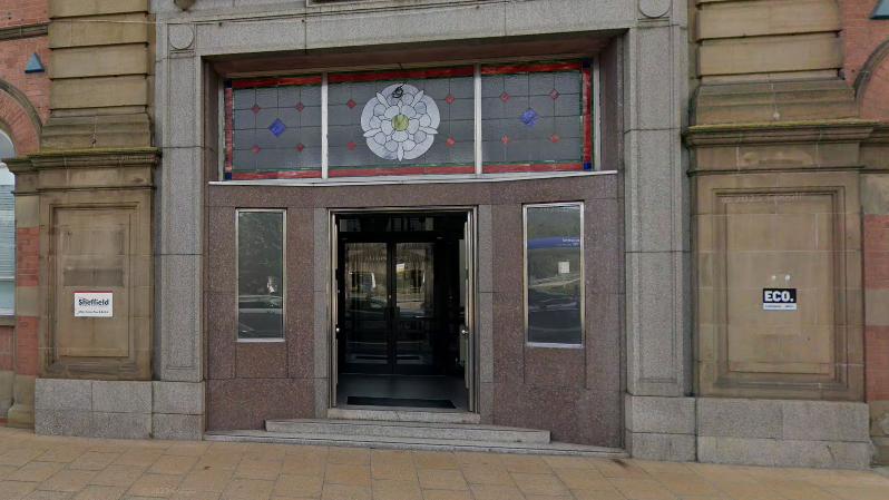 The entrance to a Victorian stone building seen from the street. A stained glass window runs horizontally above the wide doorway. In the centre window pane there is a white rose. The panes of glass either side of the centre have a pattern of blue and red diamonds.
Shallow stone steps lead up to the glass double doors in the entrance.