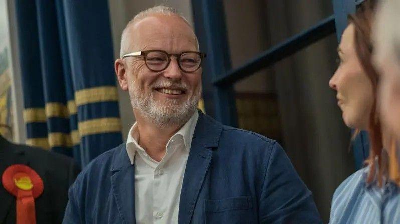 A man with short white hair and a short white beard. He wears a white shirt with the top button undone, a dark blue shirt jacket or blazer, and glasses. He is looking off camera and laughing.