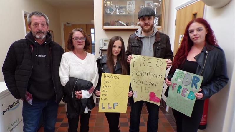 Five members of Freddie's family - his father, mother, brother and two sisters - stand holding signs saying 'For Fred', 'Fred Coleman forever 15' and 'reduce your speed and save lives'. They're all looking at the camera.