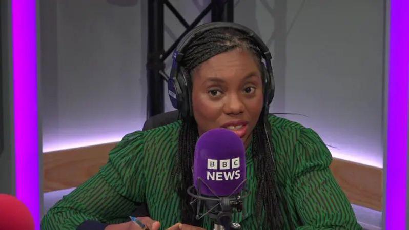 Kemi Badenoch is sitting in a radio studio wearing headphones and speaking into a purple microphone with BBC News in white writing.