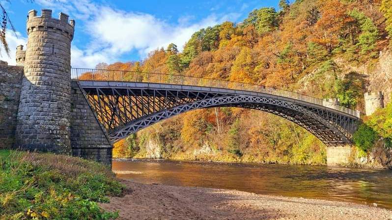 Craigellachie Bridge over the River Spey in Moray. The bridge has two stone towers with turrets at either end in a light coloured stone. The bridge is made of cast iron and has dark coloured struts coming from the top to the bottom. An arc of lighter coloured crossed struts goes from one end to the other. the sky is blue with a few thick white clouds and the landscape behind is filled with trees with a mixture of orange, brown and green colours.