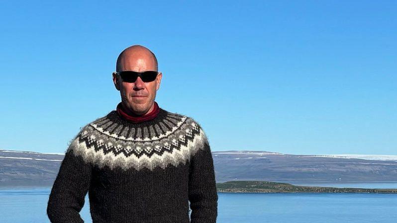 Mark Graham is standing in front of a picturesque lake. He has a shaved head and is wearing sunglasses and a grey woolly jumper.