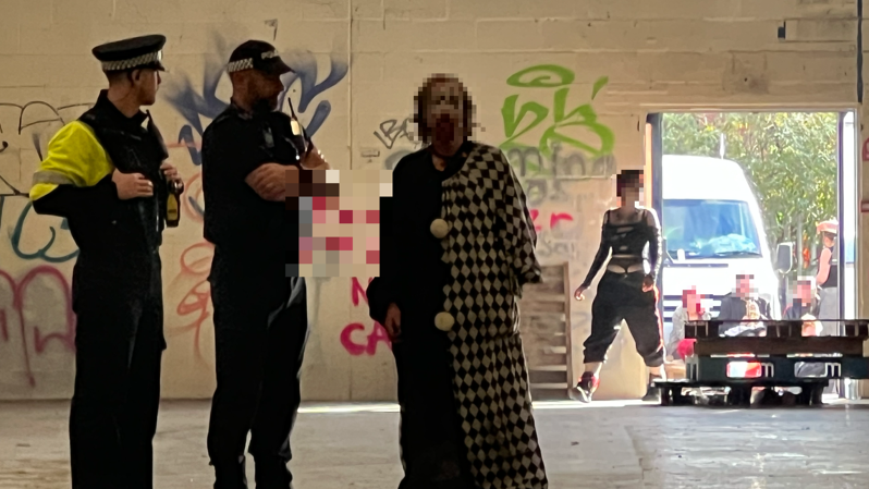 Two police officers stand in a warehouse next to a man in a clown costume. A group of people can be seen sitting down in the background. Their faces have been pixelated.