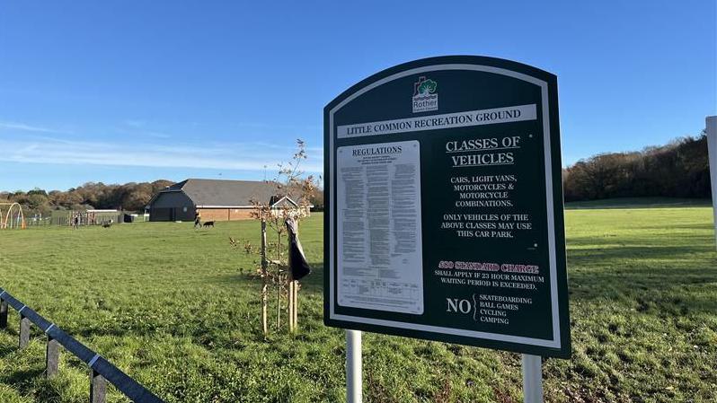 The sign for the recreation ground car park in the foreground with terms and conditions on it. In the background is the park and sports centre which cricket and football teams use