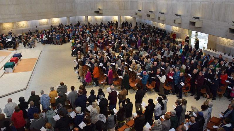 A large group of worshippers are seen inside Clifton Cathedral in Bristol during a mass. They are standing and holding orders of service and the picture is taken from above