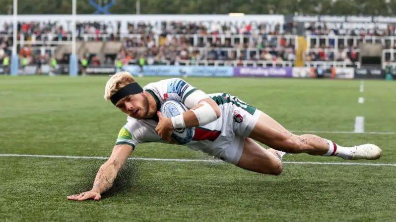 A rugby player crossing the try line and sliding forwards to score in front of fans