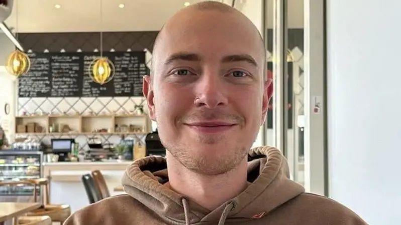 Eli smiles at the camera in a cafe. He has very short haven hair, light short stubble and dark eyebrows. He wears a brown hoodie, and has a nose ring.