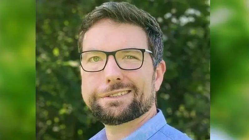 Dyn gyda gwallt brown, sbectol du a barf brown yn gwenu wrth edrych at y camera. Mae'n gwisgo crys glas.