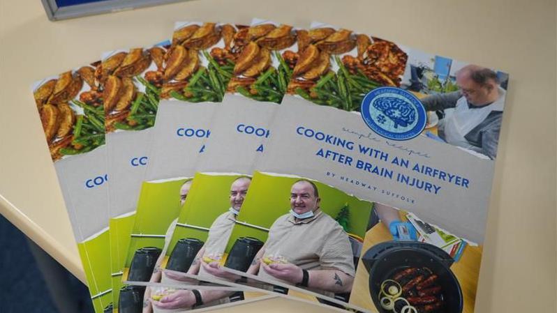 A selection of air fryer cook books rest on a table. The book title reads: "Cooking With An Air Fryer After Brain Injury." A gentleman is pictured on the book front along with an air fryer with some food placed inside it.