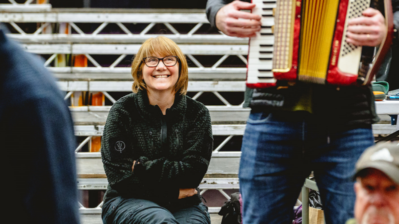 A woman with long blonde hair, glasses, a green jumper and blue trousers, sits on a step and smiles as she watches someone playing an accordion.