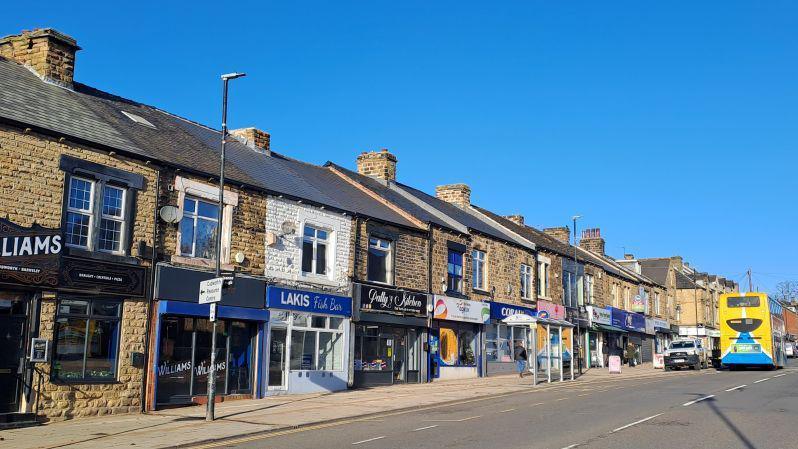 Cudworth high street is lined with traditional stone buildings. These buildings have slate roofs and chimneys. The ground floors of the buildings are occupied by various shops and businesses, each with distinct signage. Some of the visible shop signs include “WILLIAMS” and “LAKIS Fish Bar”. The shopfronts feature large glass windows and colourful signage.