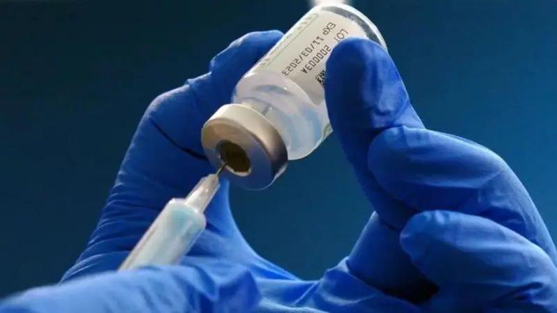 A pair of hands in blue rubber gloves pushing the needle of a syringe into a vial of vaccine.