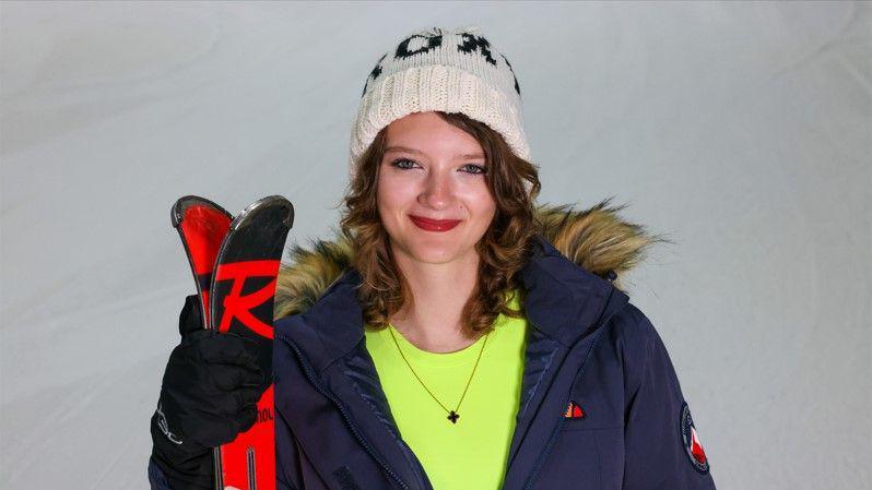 A woman on a ski slope, holding her skiis while wearing a blue coat, white hat, and neon green top