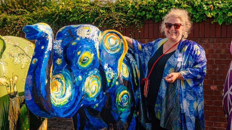 An elephant painted in the design of Vincent Van Gogh's The Starry Night, next to a blonde woman with her hand on the elephant, wearing a Starry Night cardigan