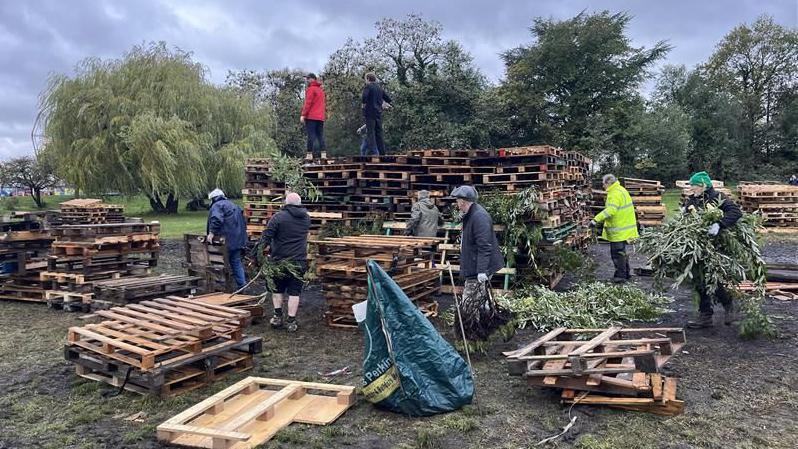 Seven people stacking large wooden pallets in a field with trees in the background.
