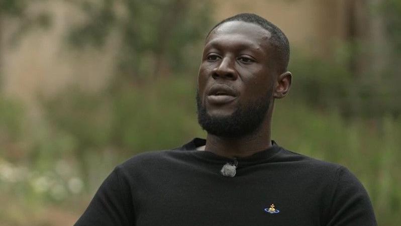 Rapper Stormzy wearing a long-sleeved black T-shirt sits in the grounds of Cambridge University, surrounded by flowers and shrubbery.